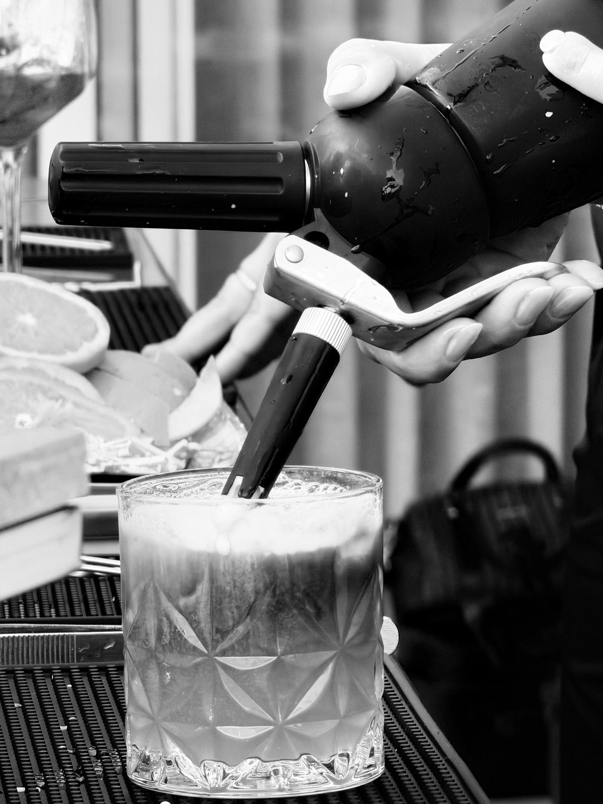 Em preto e branco, a mão do bartender finalizando um drink com o sifão